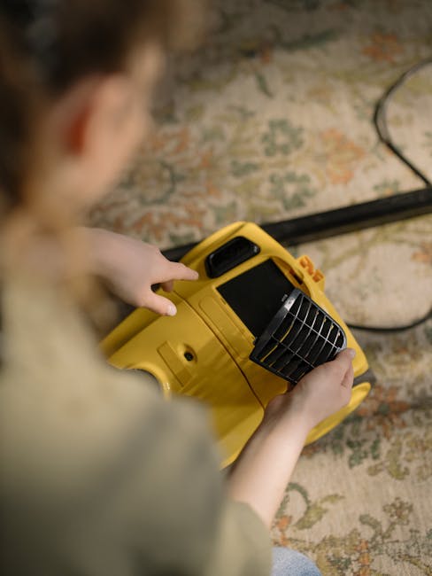 A person using a yellow and black vacuum cleaner on a patterned residential carpet in a well-lit room. The individual is wearing a green shirt and blue jeans, and their hands are placing or retrieving