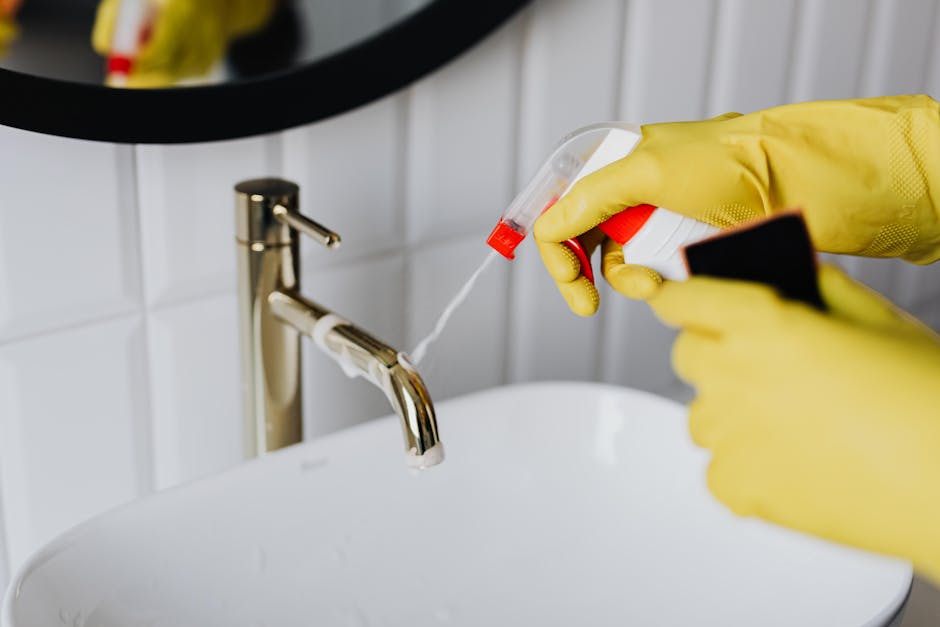 Close-up of a person's hand holding a spray bottle and applying a cleaning solution onto a dirty stove's gas hob in a kitchen setting. The stove has visible rust and grime around the burners and metal