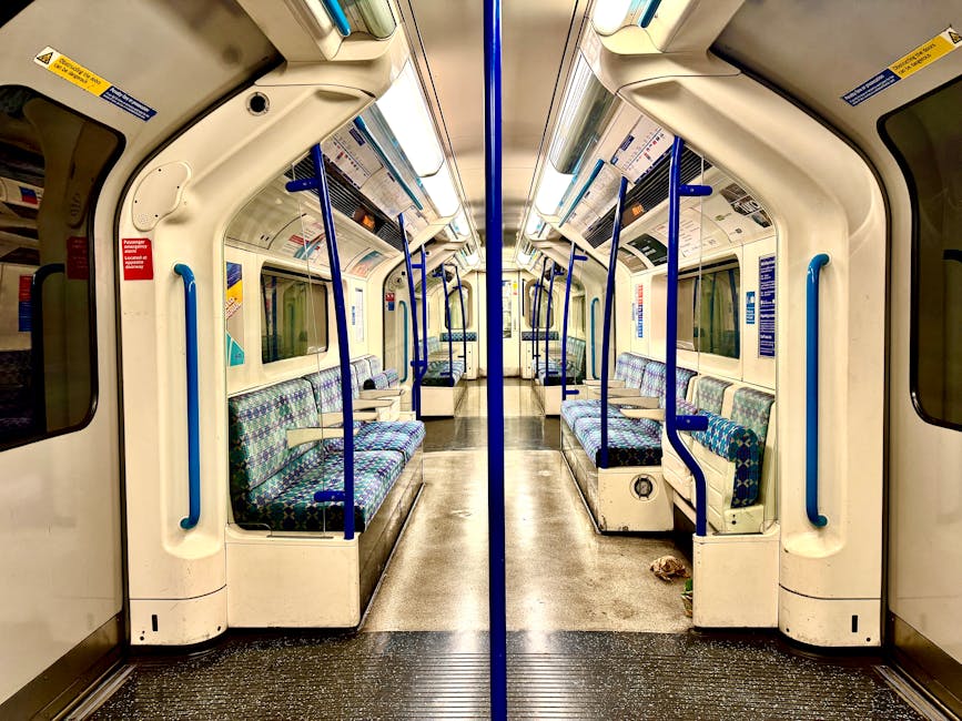 Interior of an empty commuter train carriage featuring rows of black fabric seats with a multicolored dotted pattern, arranged in a forward-facing and side-facing configuration. The carriage has a cle