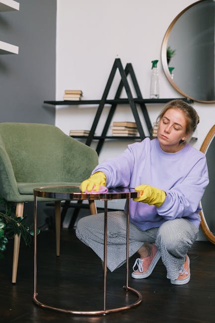 A young woman wearing a lavender sweater, yellow cleaning gloves, and grey checkered pants is kneeling on a dark wooden floor, actively wiping a small, round, copper-colored side table with a pink clo